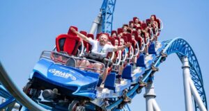 Rob Beckett discovers Europa-Park – Germany’s Largest Theme Park People riding a blue roller coaster with red seats, arms raised, against a clear blue sky, smiling and laughing as they zoom along the track.