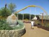 Serpent’s Curse Construction Update: How Paultons Park’s 2027 Water Coaster Is Taking Shape Amusement park path beside a stone planter with greenery, a yellow roller coaster track arching overhead, and a person walking away in a white shirt and shorts under a clear blue sky.