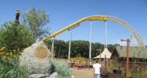 Serpent’s Curse Construction Update: How Paultons Park’s 2027 Water Coaster Is Taking Shape Amusement park path beside a stone planter with greenery, a yellow roller coaster track arching overhead, and a person walking away in a white shirt and shorts under a clear blue sky.