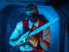 Everything We Know About Fright Nights 2026 So Far Man wearing a dark face mask and red safety vest, gripping a chainsaw in a blue-lit industrial hallway toward the camera.