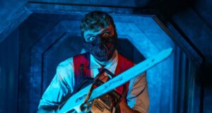 Everything We Know About Fright Nights 2026 So Far Man wearing a dark face mask and red safety vest, gripping a chainsaw in a blue-lit industrial hallway toward the camera.