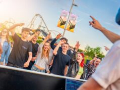 Thorpe Park Lates Starts Tonight – Everything You Need to Know Group of teens and young adults cheering and smiling at an outdoor event near a roller coaster, with a DJ on the right wearing headphones in the foreground.