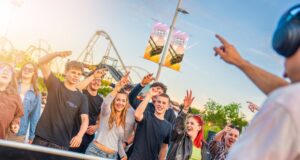 Thorpe Park Lates Starts Tonight – Everything You Need to Know Group of teens and young adults cheering and smiling at an outdoor event near a roller coaster, with a DJ on the right wearing headphones in the foreground.