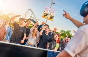 Thorpe Park Lates Starts Tonight – Everything You Need to Know Group of teens and young adults cheering and smiling at an outdoor event near a roller coaster, with a DJ on the right wearing headphones in the foreground.