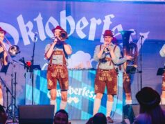 Why It’s Great to See Oktoberfest Return to a UK Theme Park Brass band performing on a stage in lederhosen; blue Oktoberfest backdrop with the word Oktoberfest behind them.