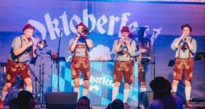 Why It’s Great to See Oktoberfest Return to a UK Theme Park Brass band performing on a stage in lederhosen; blue Oktoberfest backdrop with the word Oktoberfest behind them.